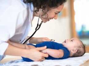 Bebé acostado en cama mientras la doctora le revisa y sonríe.