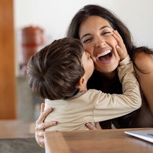 Niño dando un beso en el cachete a su madre sonriente. La mamá está sentada en frente de un ordenador portátil