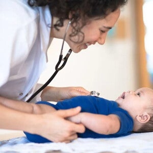 Bebé acostado en cama mientras la doctora le revisa y sonríe.