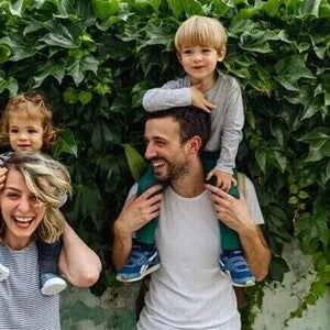 Dos adultos con dos niños sobre sus hombros, frente a una pared cubierta de hojas