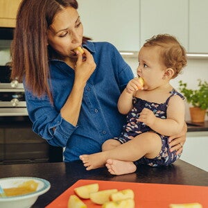 Identifica cuáles son las frutas ideales para iniciar esta nueva etapa de tu bebé. Mamá y bebé comiendo fruta en la cocina.