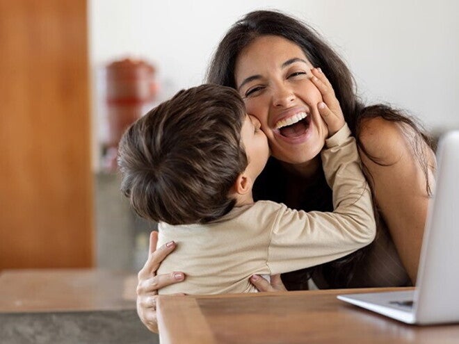 Establecer límites saludables Niño dando un beso en el cachete a su madre sonriente. La mamá está sentada en frente de un ordenador portátil