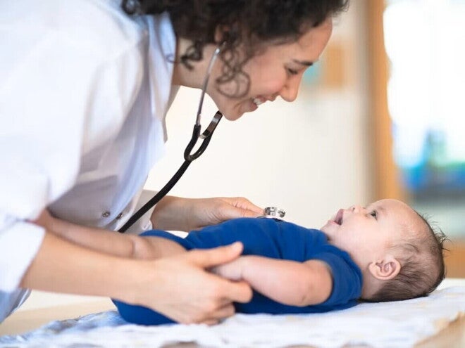 Bebé acostado en cama mientras la doctora le revisa y sonríe.