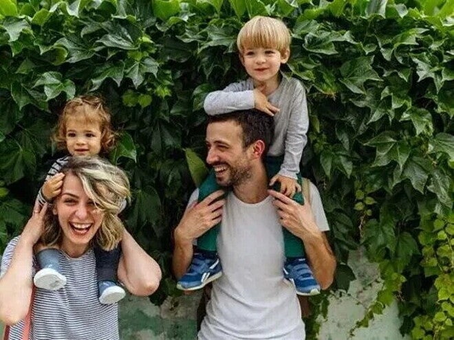 Dos adultos con dos niños sobre sus hombros, frente a una pared cubierta de hojas