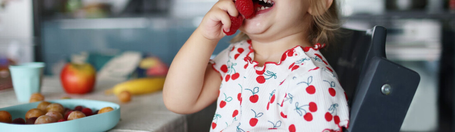 Niña pequeña en la cocina sonriendo y jugando con frambuesas en los dedos.