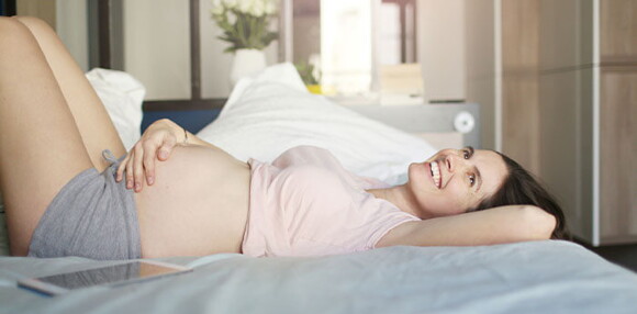 Conoce cuáles son las funciones y beneficios que tiene el líquido amniótico. Mujer embarazada sonriendo acostada en la cama tocando su vientre.