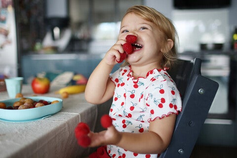 Niña pequeña en la cocina sonriendo y jugando con frambuesas en los dedos.